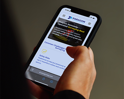 Mano sosteniendo un teléfono móvil que muestra una aplicación o sitio web con contenido informativo y elementos de interfaz en pantalla.