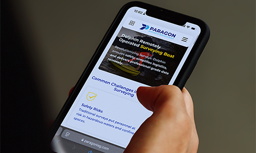 Mano sosteniendo un teléfono móvil con una aplicación o sitio web abierto, mostrando texto informativo y elementos de interfaz.